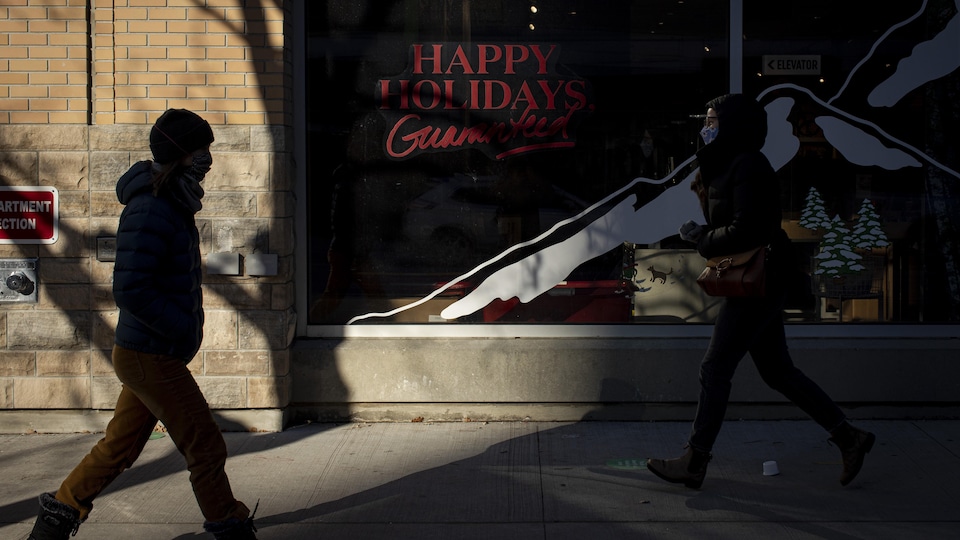 Deux passants portant des masques marchent dans les rues de Toronto devant une devanture de magasin souhaitant un bon temps des Fêtes.