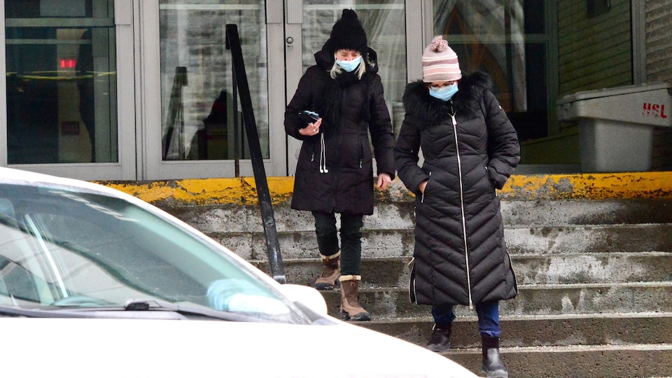 Deux femmes portant des vêtements d'hiver et des masques faciaux descendent l'escalier d'un immeuble.
