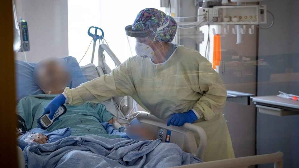 Une infirmière portant masque et visière prend les signes vitaux d'un patient dans un lit d'hôpital.
