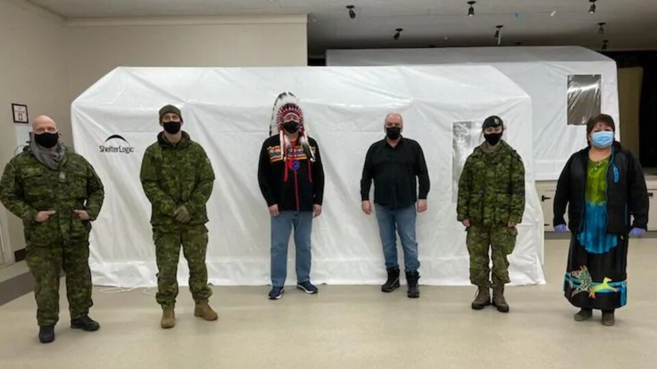 Des militaires et des Autochtones qui portent un masque devant une tente blanche.