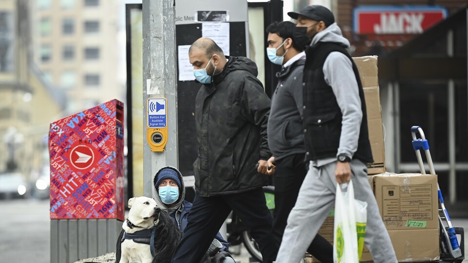 Trois hommes portant un masque passent dans la rue à côté d'un mendiant assis au sol, qui porte lui aussi un masque.