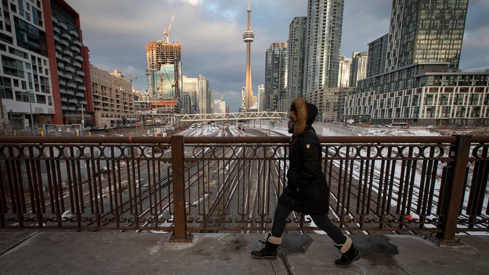 Une Torontoise marche dans la rue avec un masque; la Tour CN est en arrière-plan.