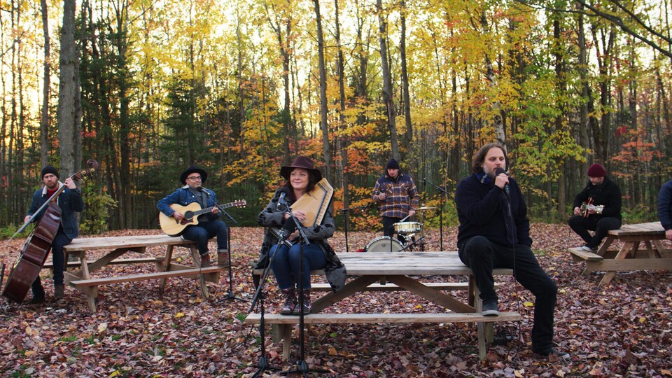 Des musiciens et une musicienne tenant chacun leur instrument chantent, assis dans un boisé en automne.
