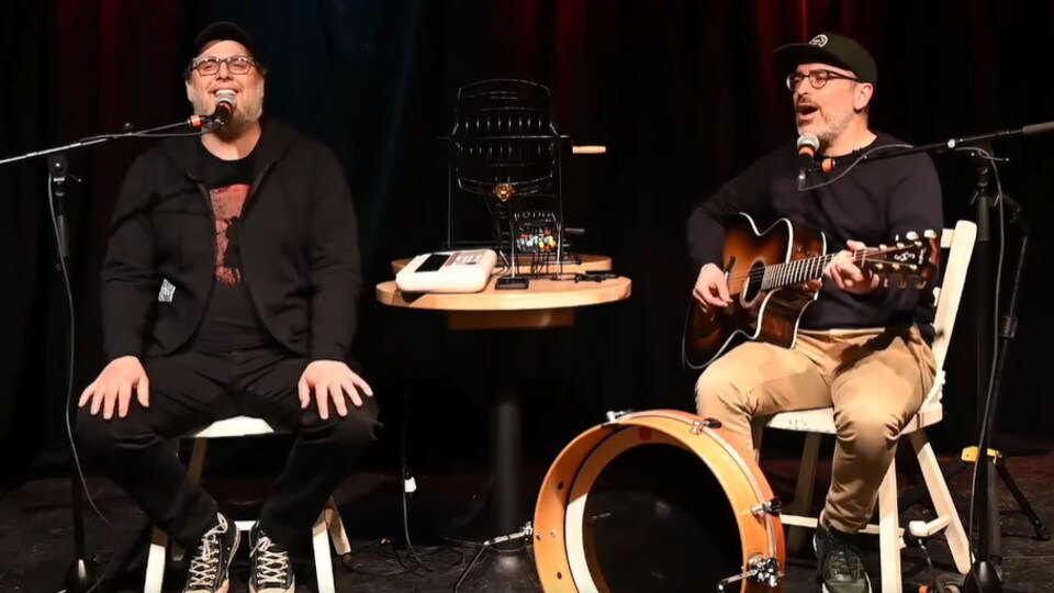 Les deux hommes interprètent une chanson assis sur des tabourets. 