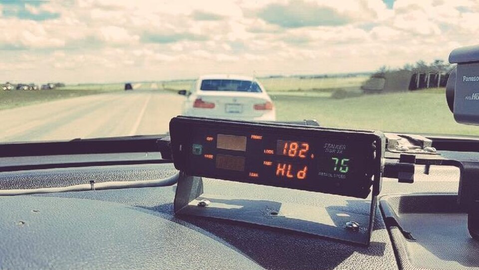 Vue depuis la place conducteur de la voiture de police, le radar au premier plan indique la vitesse de 182 km/h. En arrière plan, de l'autre côté du parre-brise, se trouve une voiture blanche stationnée sur le bas-côté.