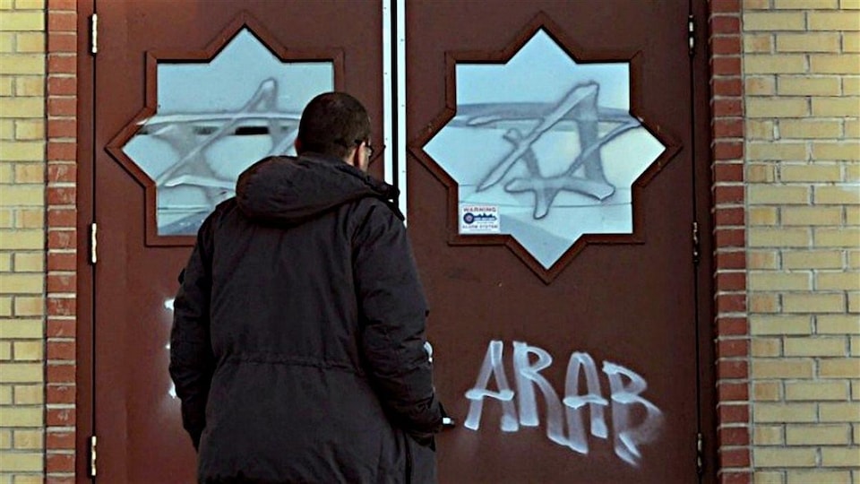 Une homme debout face à des portes fermées sur lesquelles des graffitis ont été inscrits, dont le mot « arab », en anglais.