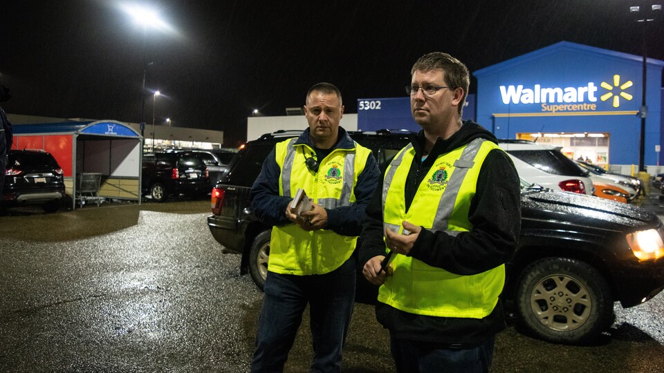 Deux hommes portant des dossards jaunes dans un stationnement d'un Walmart, le soir.