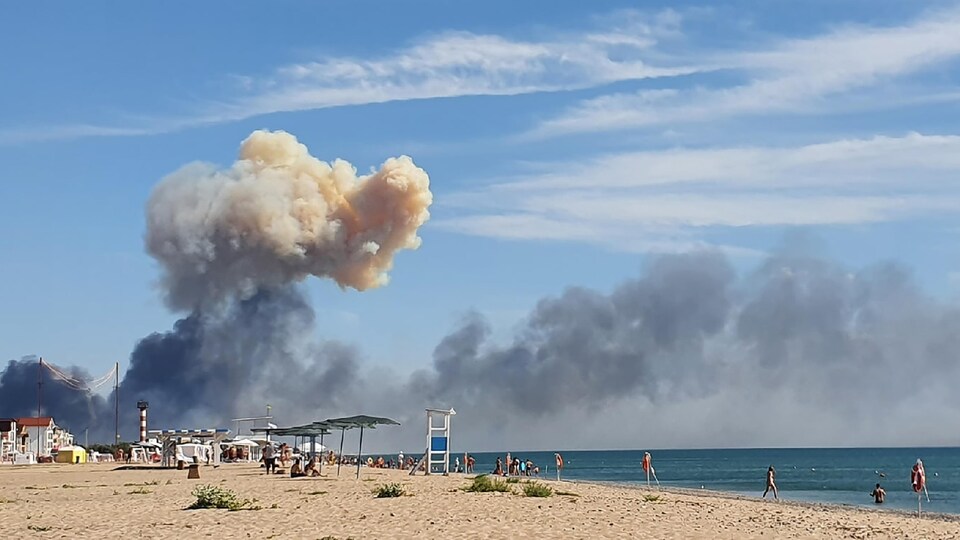 De la fumée s'élève dans le ciel, près d'une plage où des gens profitent du beau temps.