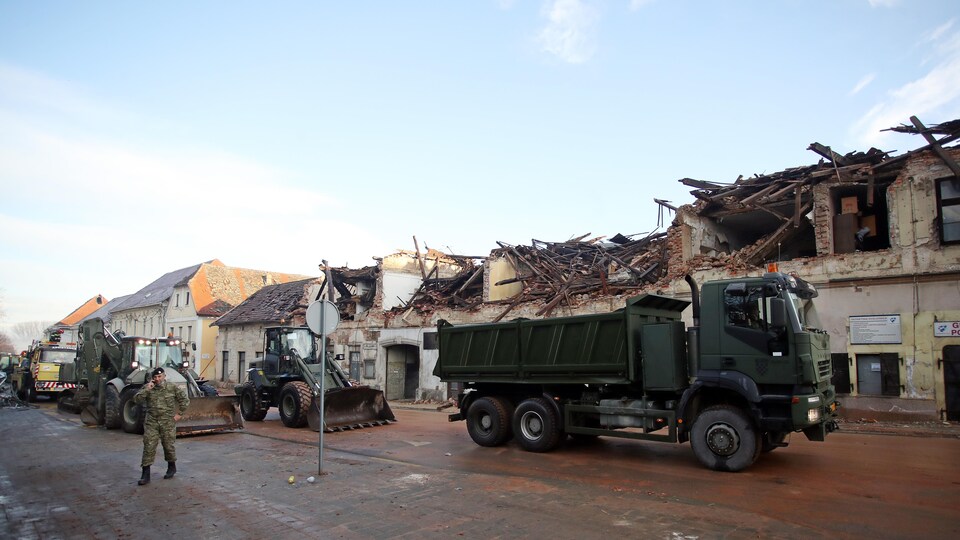 Un camion de l'armée et des excavatrices sont stationnés devant une rangée de bâtiments démolis.