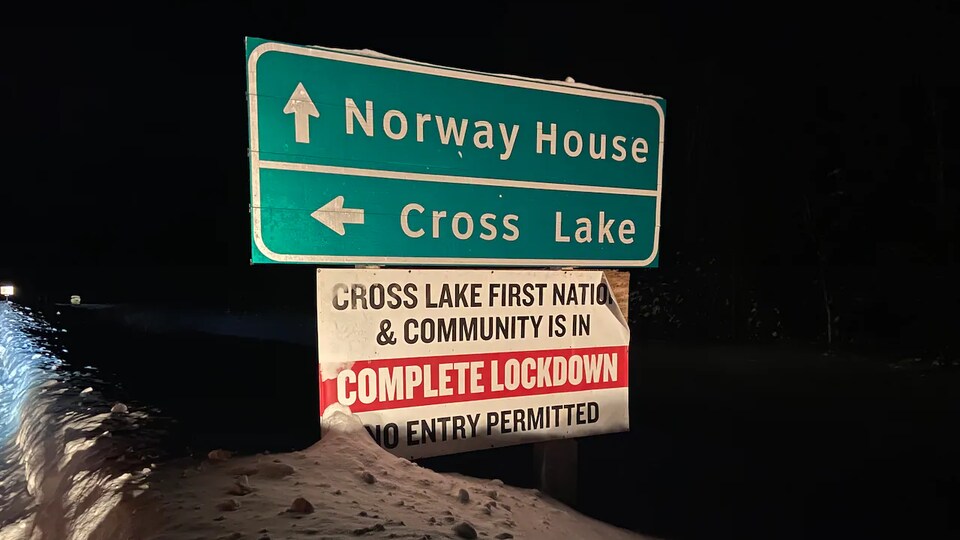 Une pancarte routière dans le noir indique les communautés de Norway House et de Cross Lake. Sous ces indications, une autre pancarte précise que la Première Nation de Cross Lake est en confinement.