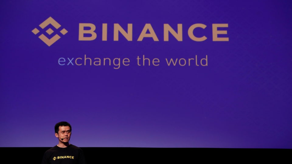 Un homme parle sur scène lors d'une conférence. Derrière lui, le logo de Binance est affiché sur un écran.