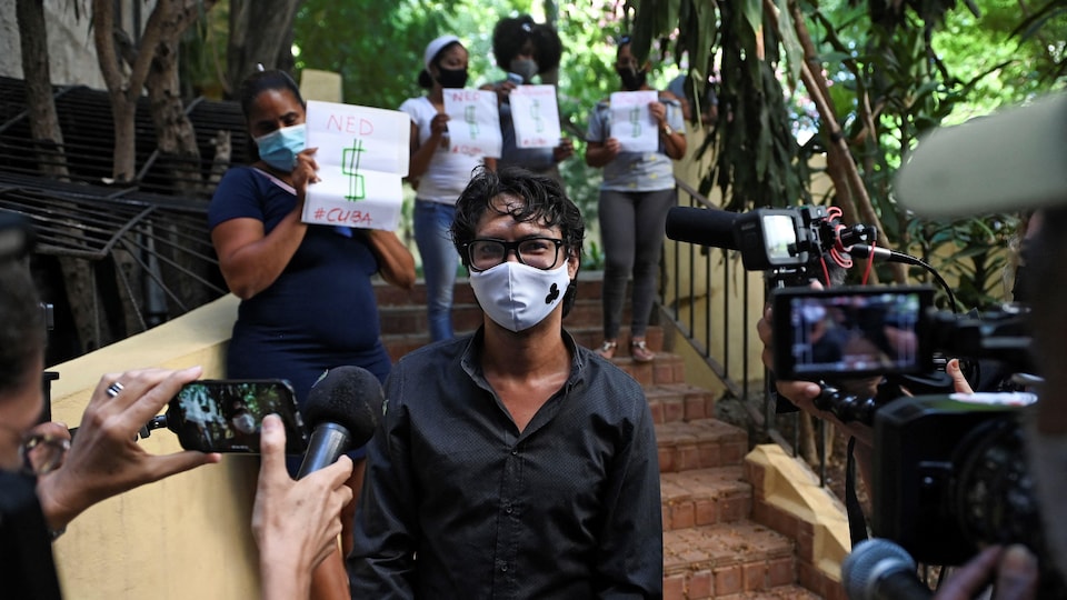 Un homme portant un masque est entouré par des journalistes; à l'arrière-plan, des femmes portent des pancartes.