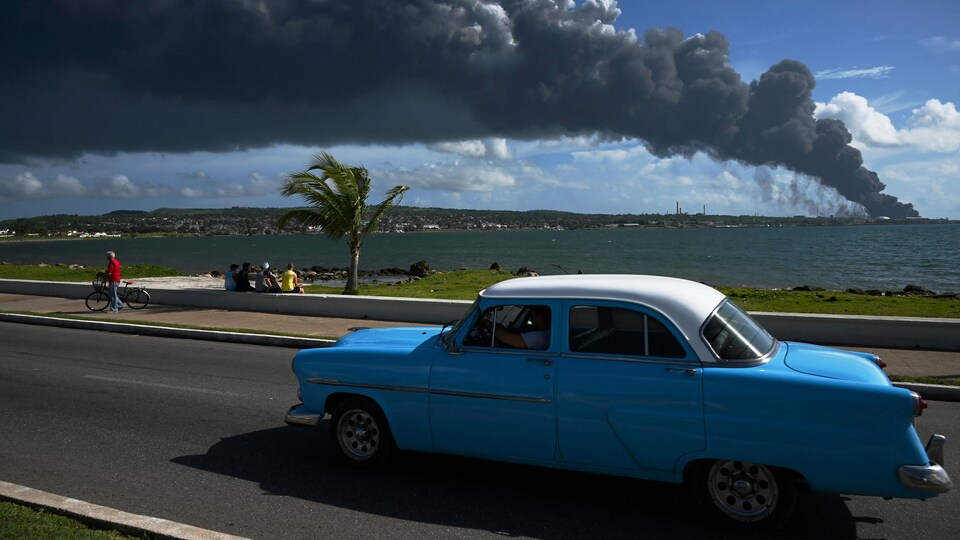 Une vieille voiture américaine passe dans une rue de Matanzas, avec en arrière-plan la fumée noire d'un réservoir de pétrole en feu.