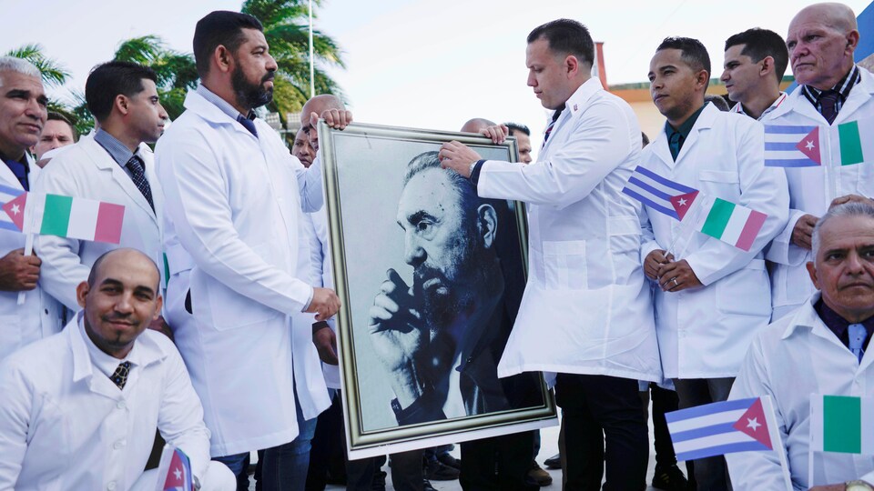 Plusieurs hommes en sarrau avec des petits drapeaux de Cuba et d'Italie, deux tiennent un portrait de Fidel Castro.