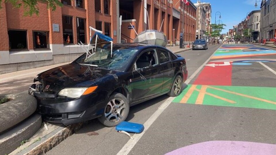Une voiture est stationnée sur la rue et elle est endommagée. Une chaise fracasse le pare-brise alors qu'un engin aérospatial a percuté l'arrière du véhicule.