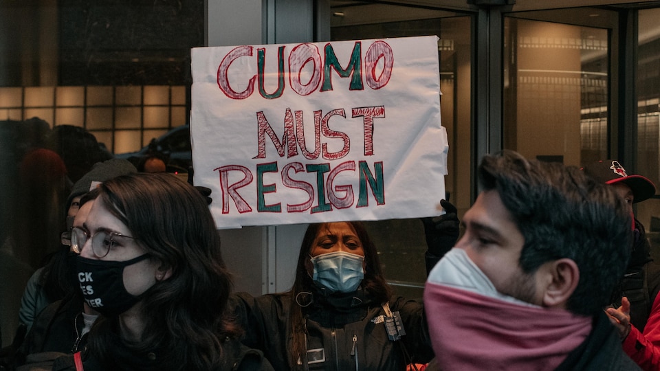 Entourée d'autres manifestants, une jeune femme tient une pancarte disant : « Cuomo doit démissionner ». 
