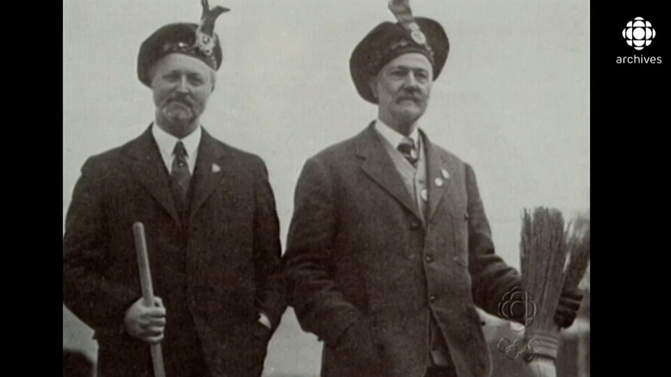 Deux hommes portant le costume traditionnel écossais et tenant chacun un balai. 