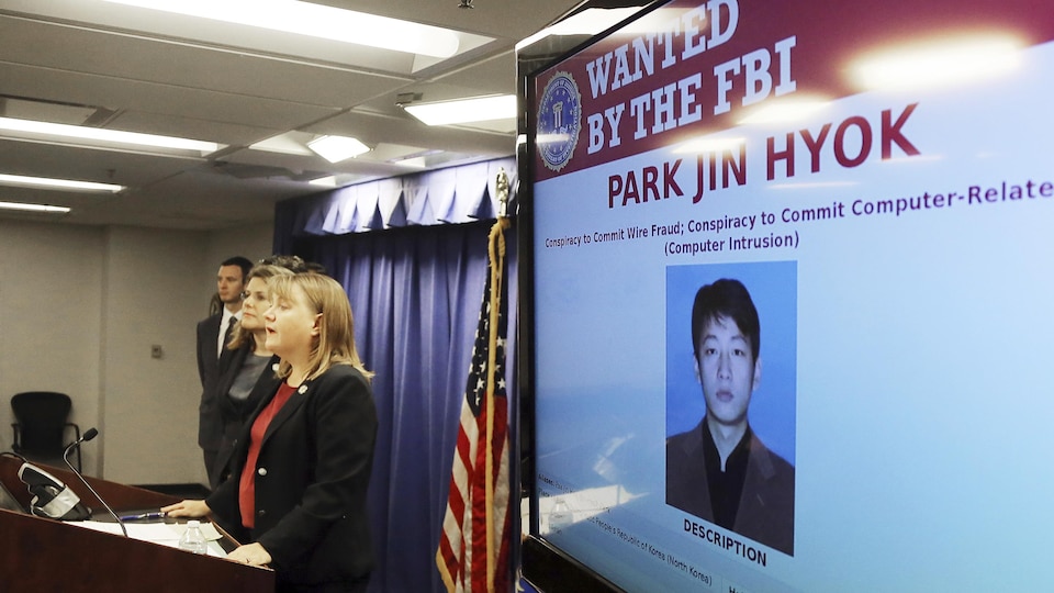 Deux femmes et un homme debout à l'avant d'une salle de conférence et, sur un écran, la photo d'un homme asiatique surmontée de son nom : Park Jin-hyok.