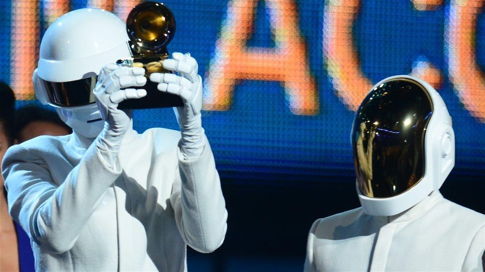 Les deux hommes sont vêtus de blanc et portent chacun un casque intégral qui leur cache le visage. Un des deux tient un trophée dans ses mains.