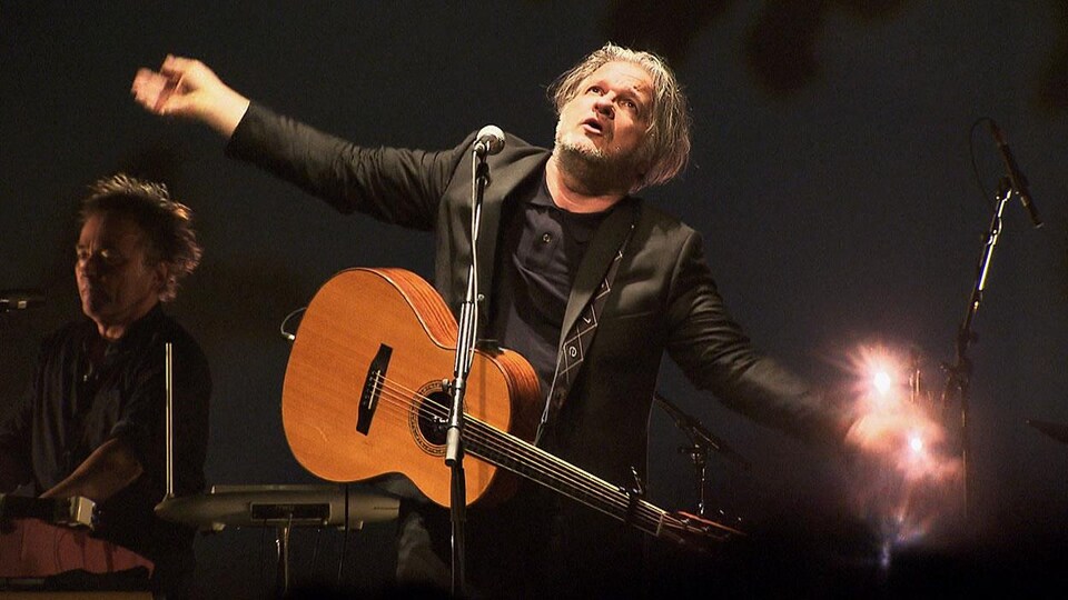 Daniel Bélanger, avec sa guitare, qui ouvre grand les bras sur scène. 