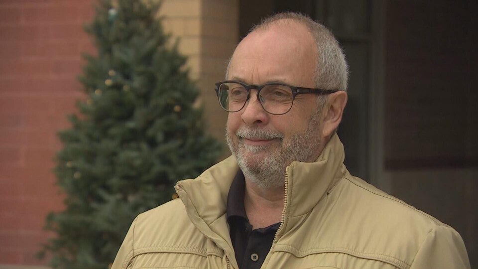 Daniel Boucher donne une entrevue à Radio-Canada à l’extérieur d’un bâtiment en briques, vêtu d’un manteau couleur ocre. Derrière lui on retrouve un sapin. 