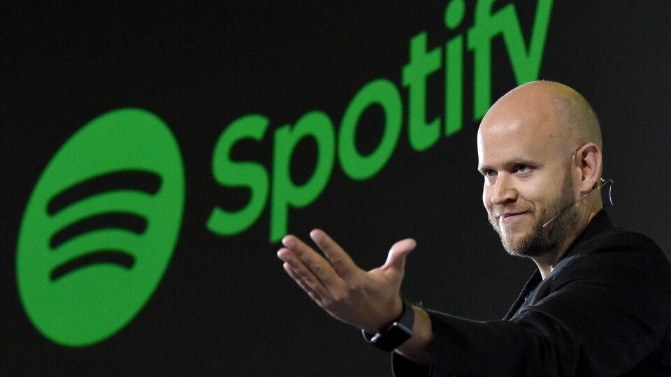 Un homme souriant, ouvre la main, devant un logo de Spotify. 