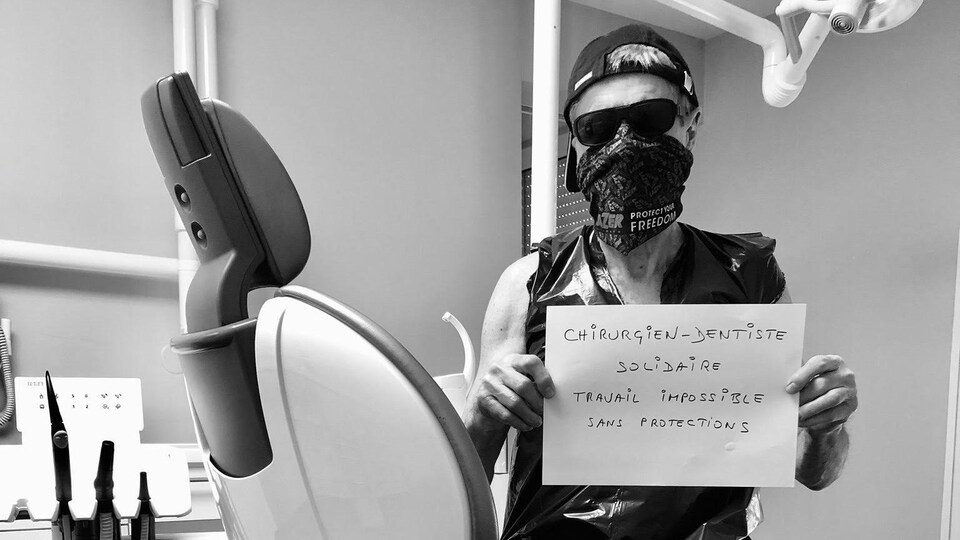 Un homme pose sur une chaise de dentiste avec une pancarte sur laquelle on peut lire : « Chirurgien-dentiste solidaire travail impossible sans protections ».