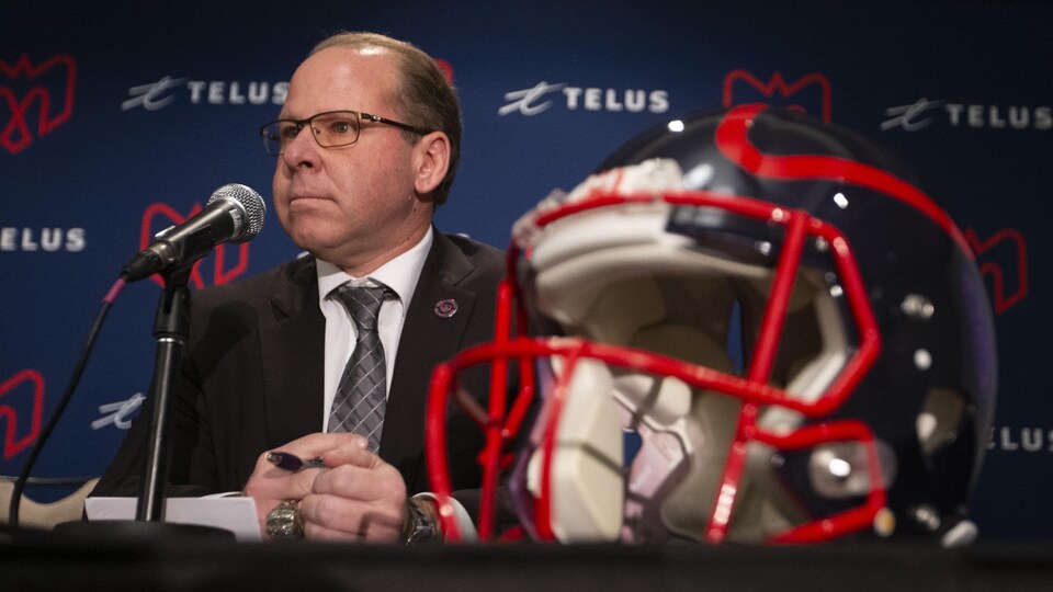 Il est assis à une table derrière un micro et un casque de football.