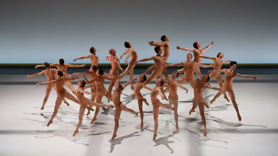 Une vingtaine de danseurs et danseuses portant des costumes beiges sautent simultanément, créant une sorte de cercle avec leur corps.