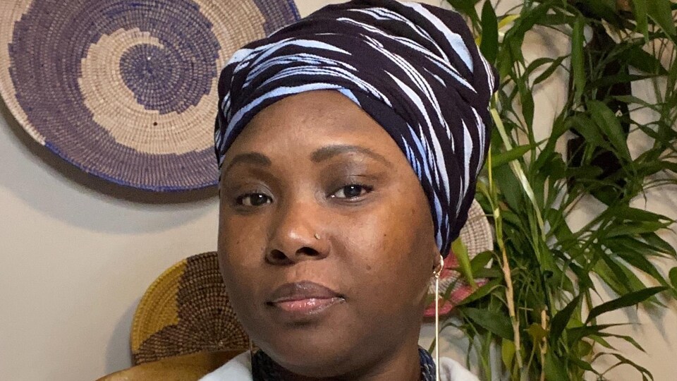 Darlene sourit et observe la caméra. Elle porte un turban cachant ses cheveux et un chemisier, un collier à son cou, des boucles d'oreilles et un perçage au nez. En arrière-plan, une plante et des paniers en osier accrochés au mur forment le décor.