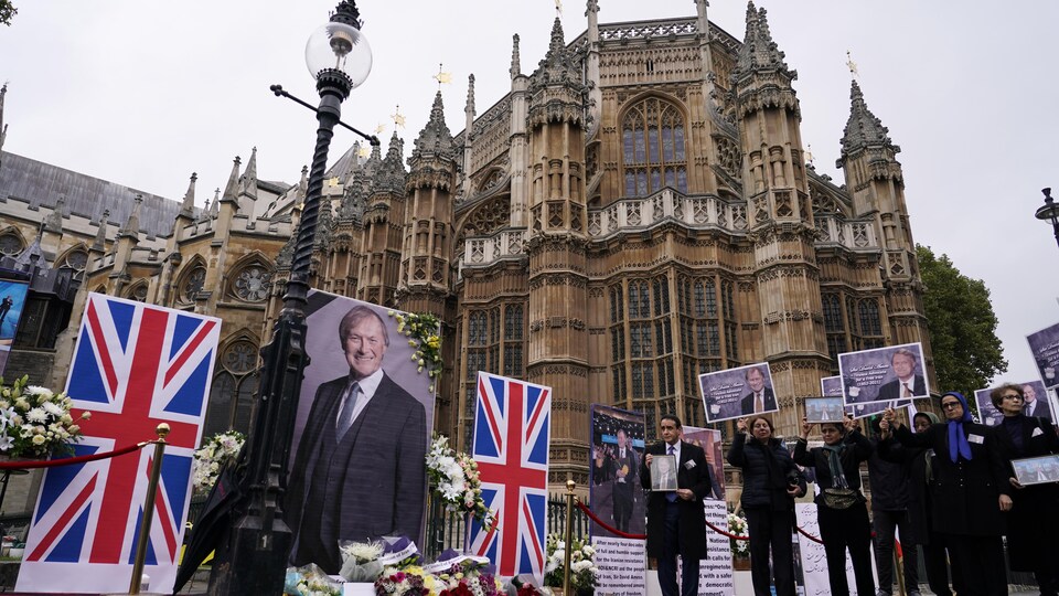 Des gens tiennent des pancartes de David Amess près du parlement. Des fleurs ont été déposées devant une grande affiche du député, installée entre deux drapeaux britanniques. 