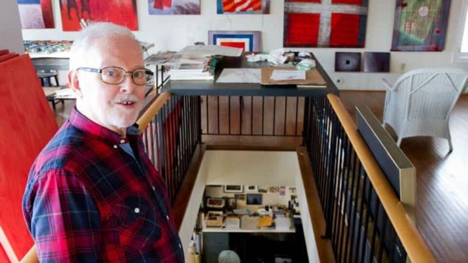 David Blackwood photographié en haut d'un escalier, dans une pièce dont les murs sont décorés de tableaux et de peintures.