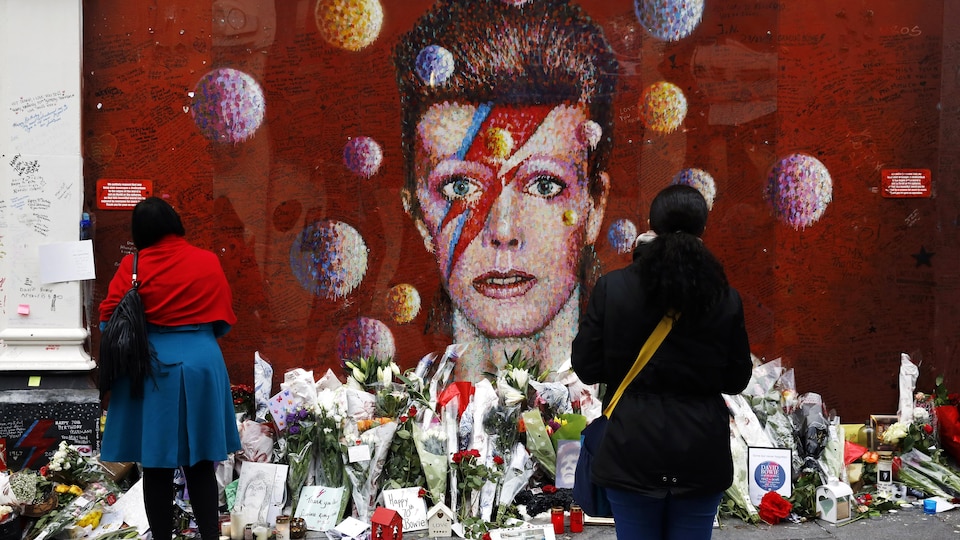 Deux personnes debout regardent un mur extérieur sur lequel est peint le visage de David Bowie sur fond rouge. Des fleurs et de nombreux messages d'amour jonchent le sol.