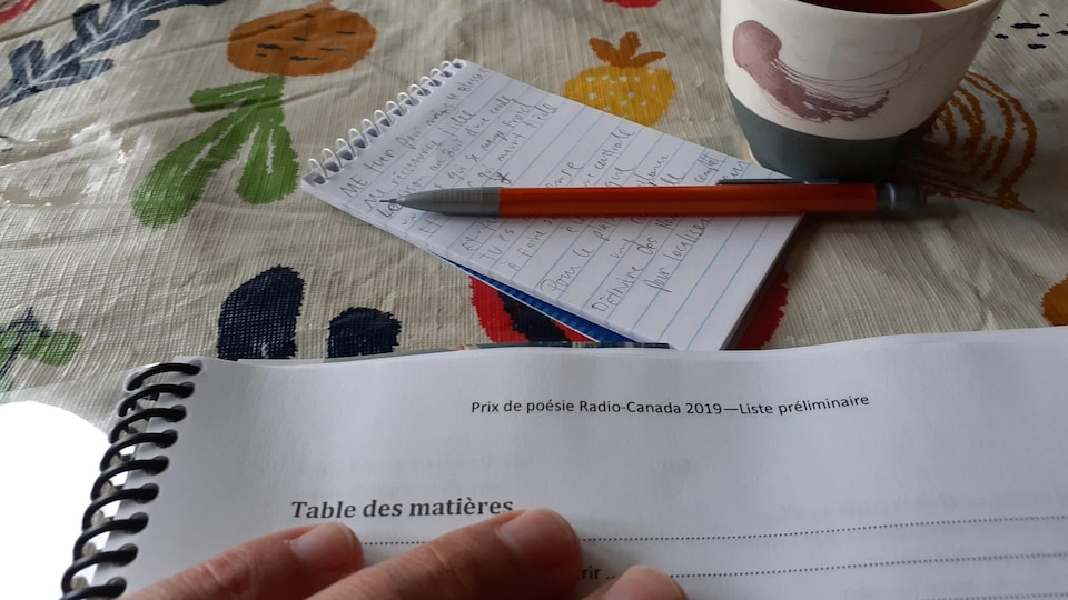 Photo en gros plan d'une main posée sur un document relié par un boudin noir, portant le titre « Prix de poésie Radio-Canada 2019, Liste préliminaire » posé sur une toile cirée fleurie. En arrière plan, un carnet de notes, un pousse-mine et une tasse de thé. 