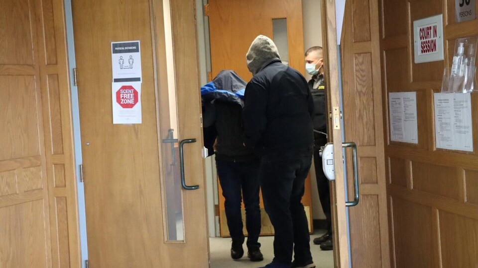 Un homme se couvre de son manteau à la sortie d'une salle d'audience. Il est entouré de deux hommes.