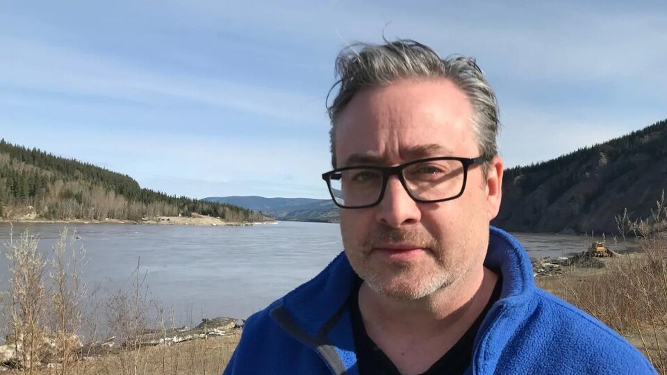 Un homme aux cheveux gris portant des lunettes devant un cours d'eau entouré de montagnes