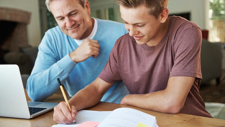 Un adolescent écrit dans un cahier, son père à ses côtés.