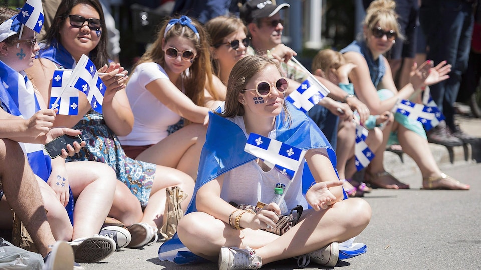 De jeunes filles brandissant des drapeaux du Québec sont assises dans la rue pour assister au défilé de la Fête nationale.