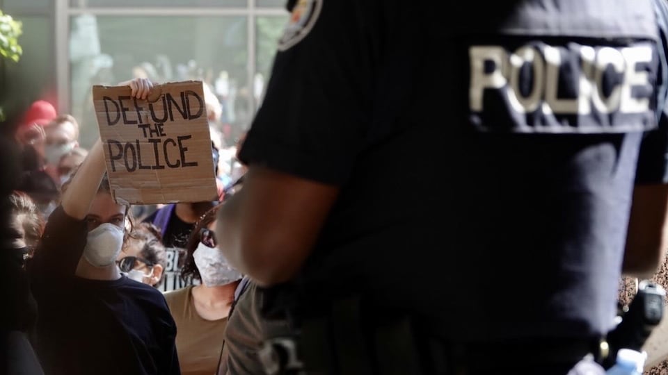 Une jeune femme brandit une pancarte où il est écrit « Defund the police » devant un agent torontois.