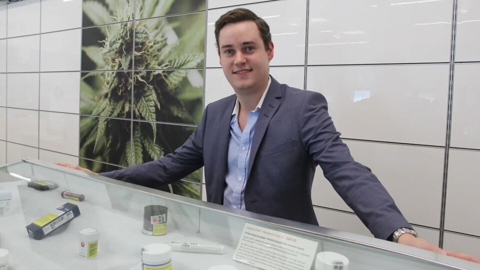 John Arbuthnot est devant un comptoir de magasin et sourit. Une grande image de fleurs et de feuilles de cannabis est affichée au mur derrière lui.