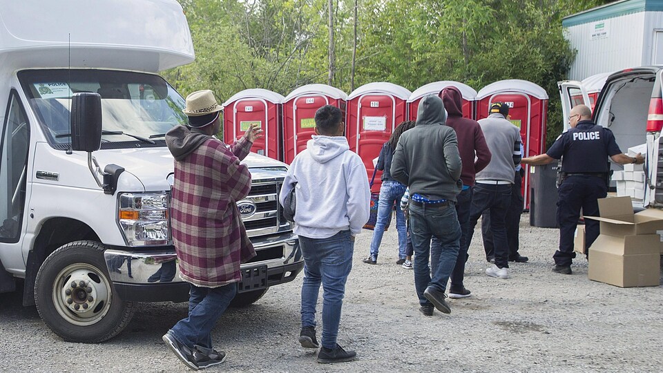 Six demandeurs d'asile arrivés par le chemin Roxham, près de Saint-Bernard-de-Lacolle, reçoivent des boîtes à lunch des mains d'un policier, alors qu'on voit des toilettes chimiques en arrière-plan.