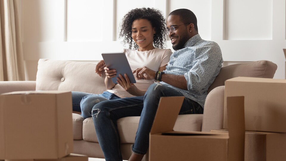Un couple en plein déménagement regarde une tablette assis sur un divan. 