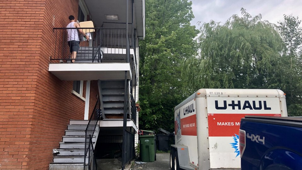 Un homme grimpe l'escalier extérieur d'un immeuble avec des boites. Une camionnette et une remorque se trouvent dans la cour.