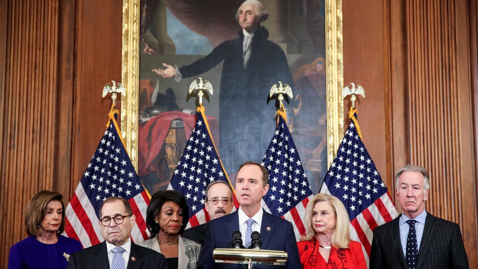 Sept dirigeants démocrates, dont Adam Schiff, qui parle devant un micro, tiennent une conférence de presse avec en arrière-plan quatre drapeaux américains et le portrait de George Washington.