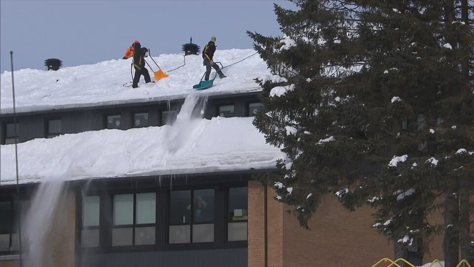 Deux hommes se trouvent sur le toit d'une école et déneigent avec des grosses pelles.
