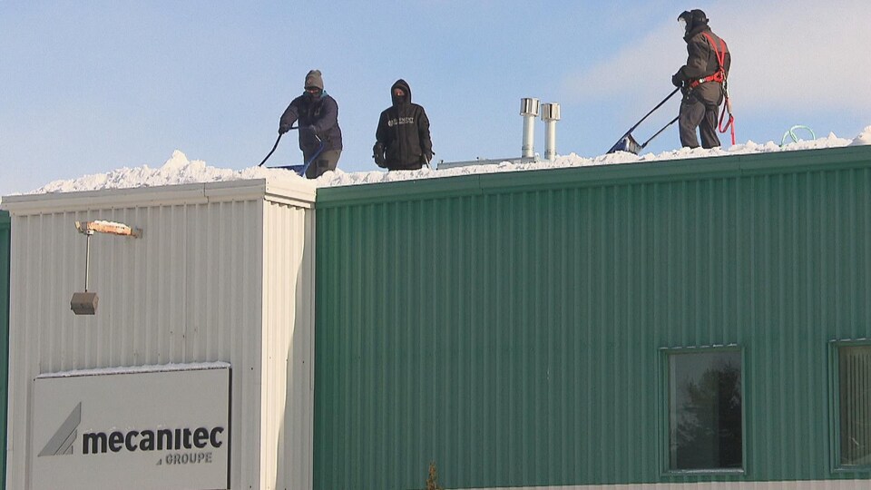 Trois hommes pellettent la neige sur la toiture d'un bâtiment commercial.