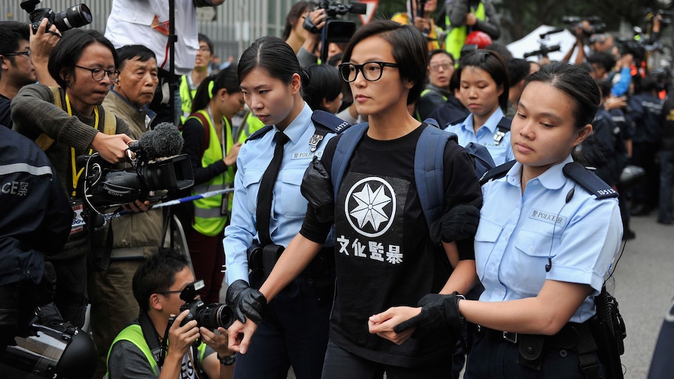 En état d'arrestation, Denise Ho est escortée par deux policières devant un groupe de journalistes, de photographes et de caméramans.