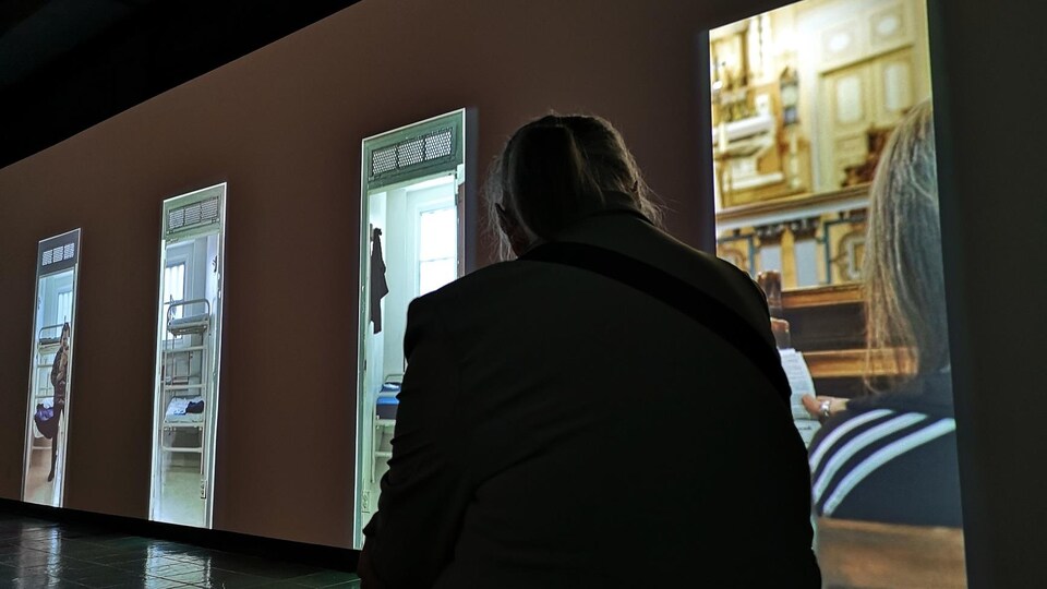 Une femme regarde des vidéos projetées sur un mur dans une salle noire. 