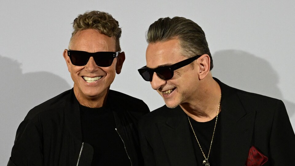 Martin Gore et Dave Gahan portent des lunettes fumées et sourient à la caméra. 