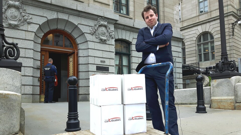 Renaud Brossard prend la pose devant l’entrée de l’édifice Honoré-Mercier. Il a les bras croisés et se tient derrière un charriot plat sur lequel ont été déposées les quatre boîtes en carton renfermant les 75 000 signatures recueillies par la pétition de la FCC.